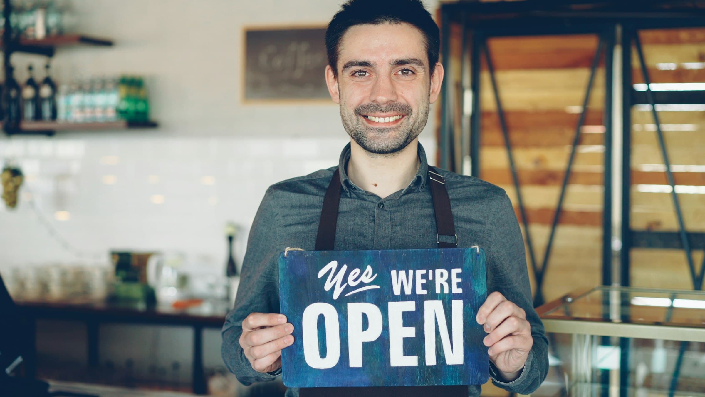 A cafe owner in an apron holds up a wooden open sign at his counter.