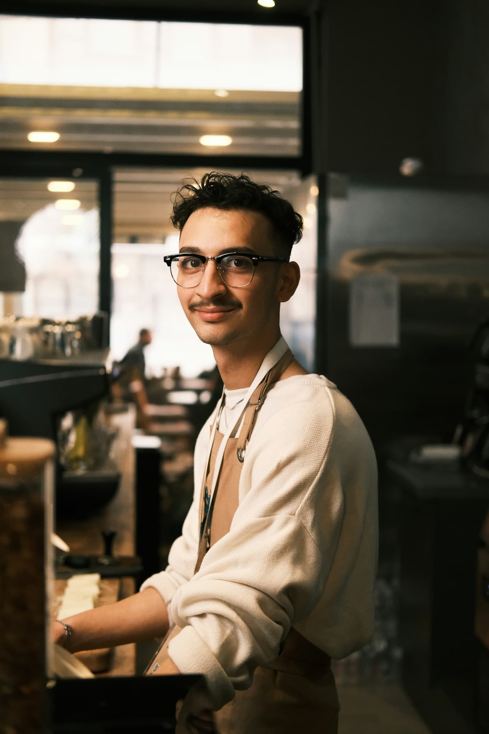 A cafe owner in an apron stands at his counter, looking at the camera with a warm smile.