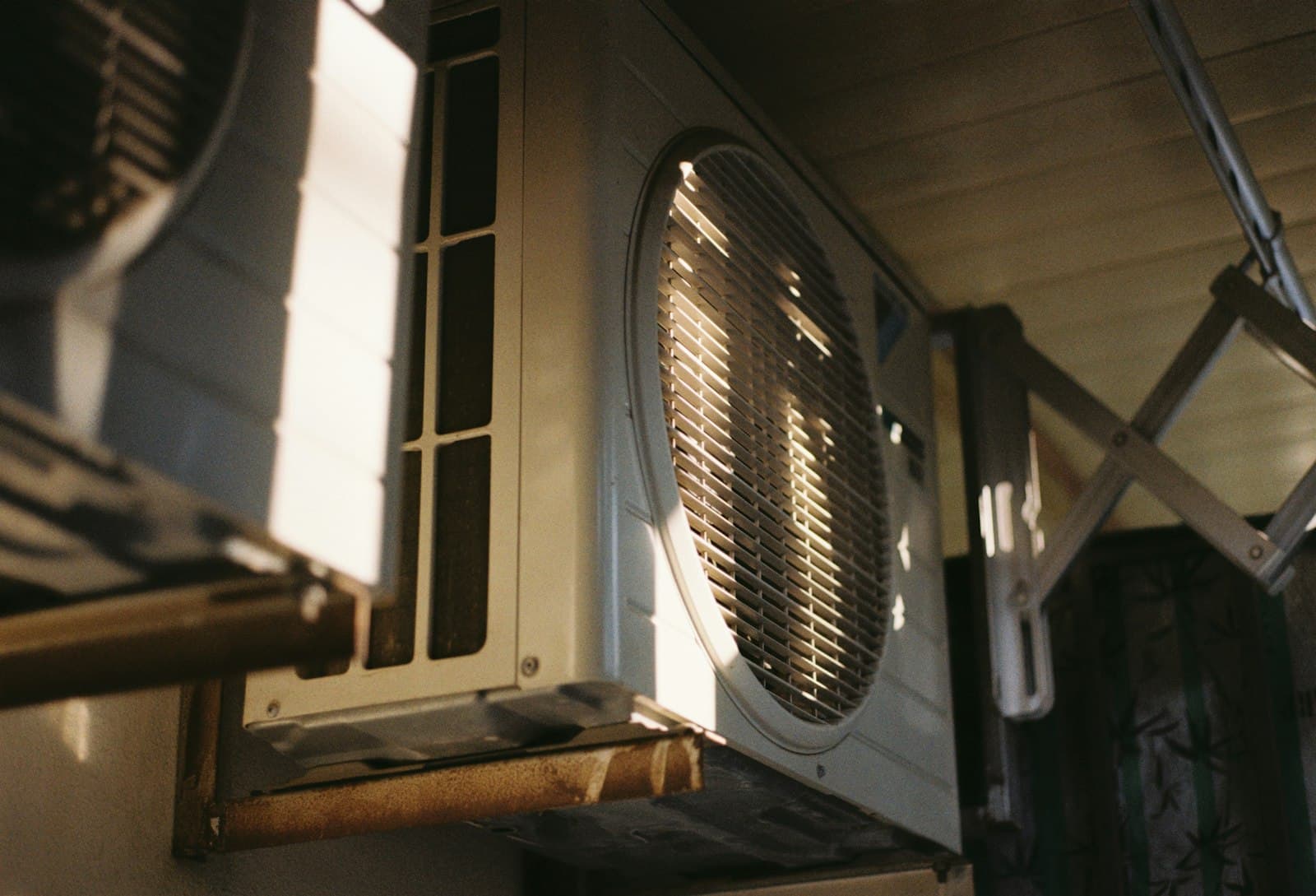 An outdoor air-conditioning condenser mounted against a wall, its circular fan grille lit by low afternoon sun.
