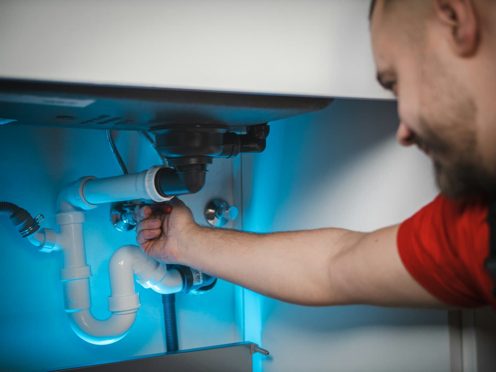 A plumber in a red shirt reaching under a sink to tighten the pipework of a drain trap.