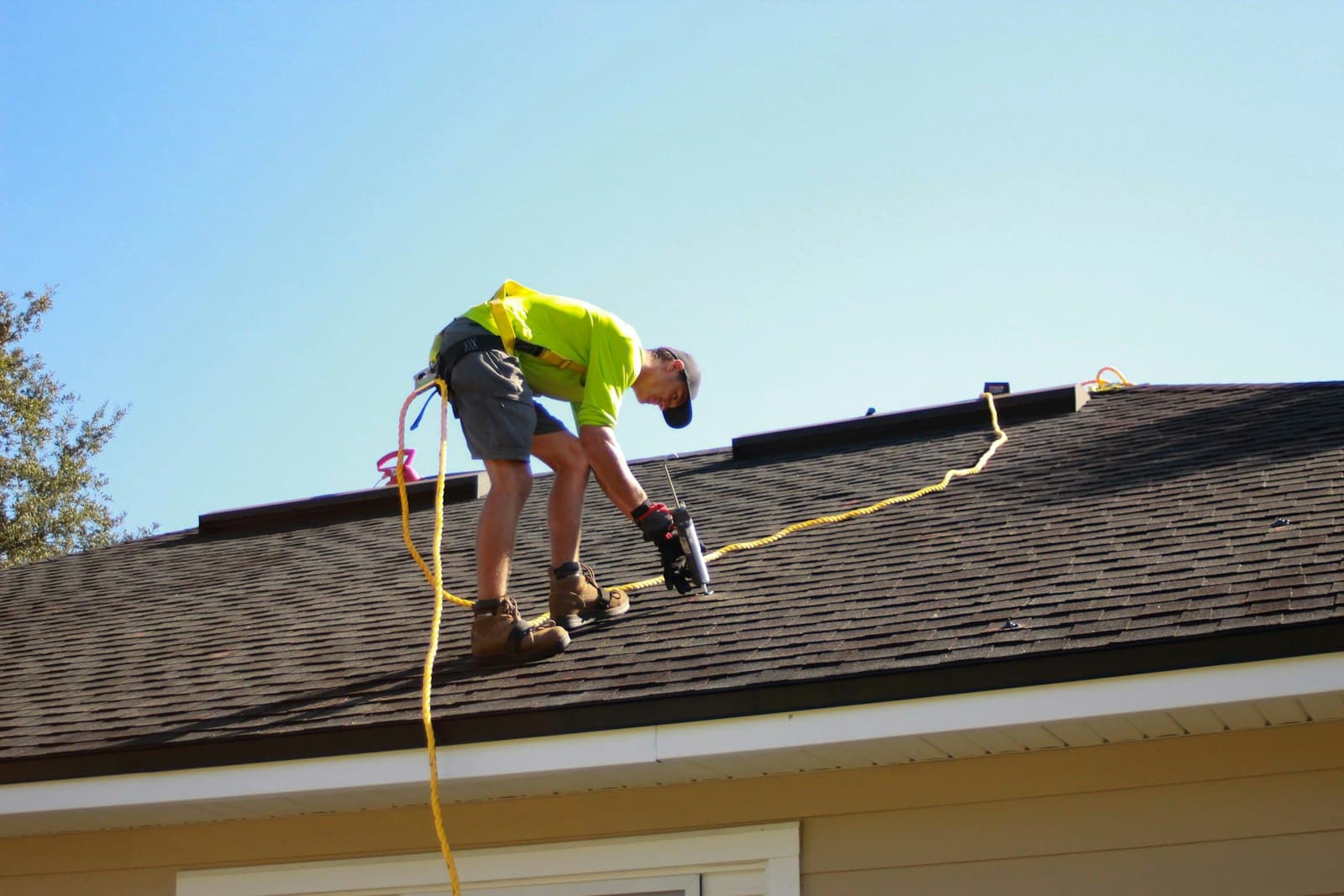 A roofer in a high-visibility shirt bent over a residential shingle roof, driving fasteners with a pneumatic nail gun, a yellow safety line running across the shingles.