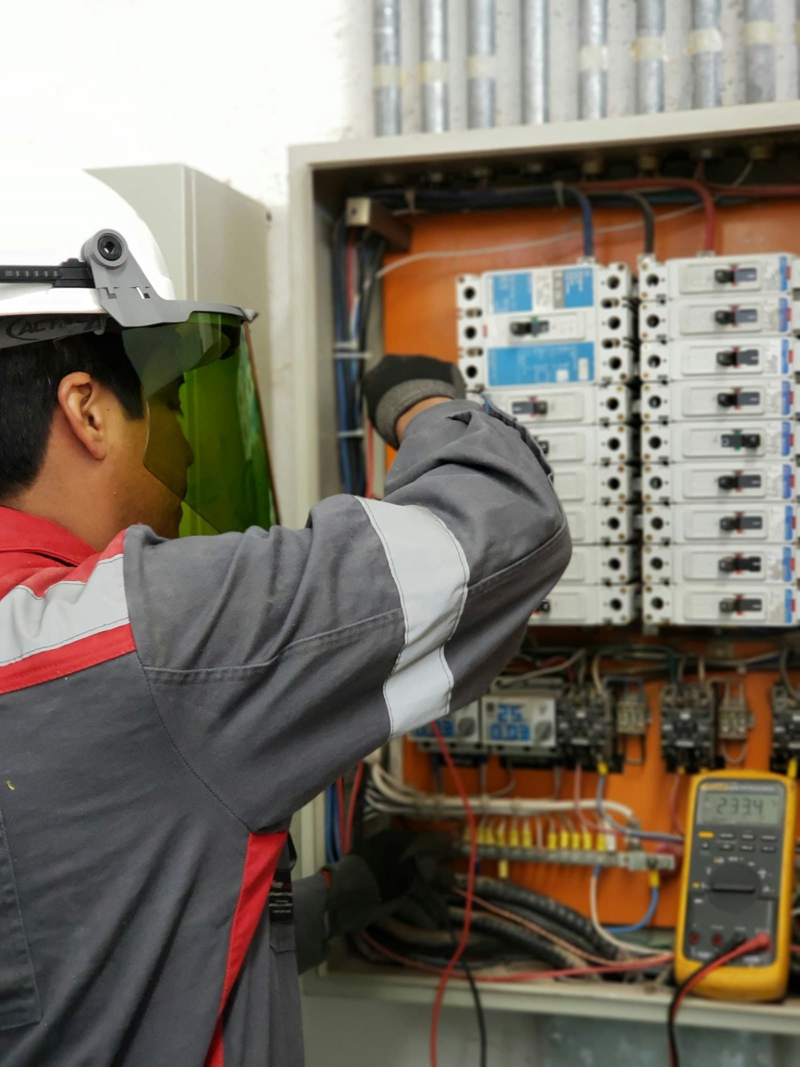 An electrician in a hard hat and green face shield reaching into an open electrical panel, testing a row of circuit breakers with a handheld meter.