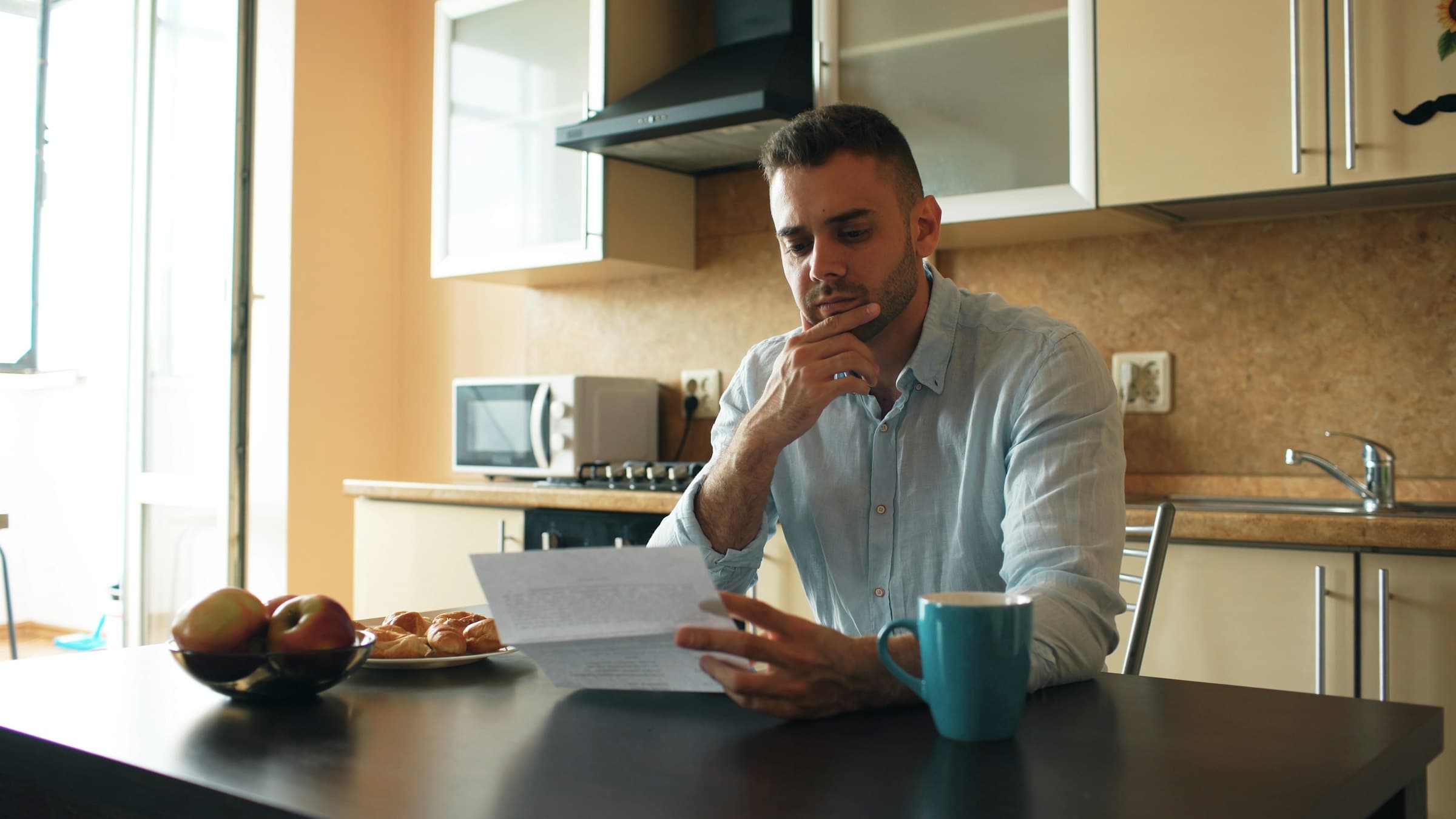 A man at a kitchen table reading a printed statement, a mug of coffee beside him.